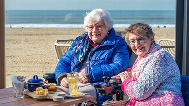 Op deze foto zie je een medewerker van de Zonnebloem aan het strand samen met een oudere persoon in een rolstoel. Dankzij rolstoelauto kan Magda uitwaaien op het strand.