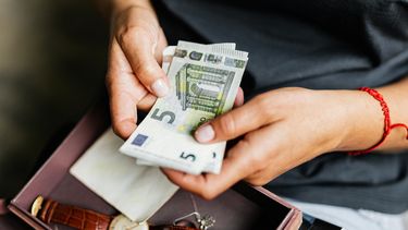 Een persoon telt vanwege inflatie vijf-eurobiljetten met beide handen. Op de achtergrond is een doos zichtbaar met een bruine leren armbandhorloge en andere kleine voorwerpen.