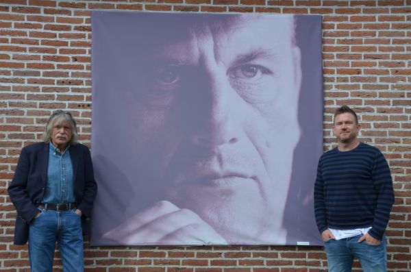 Een foto van Johnny de Mol met Johan Derksen bij een afbeelding van Harry Muskee in Grolloo