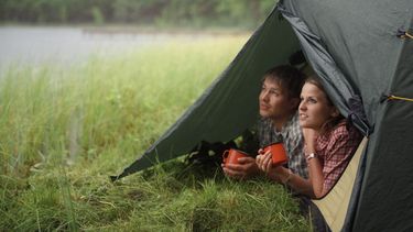 Een foto van een jong stel dat samen uit een tent kijkt op de groene oever van een meer. Ze hebben oranje kampeerbekers vast. Staatsbosbeheer sluit alle wildkampeerterreinen.