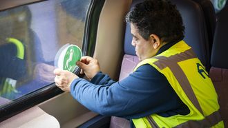 Een foto van een NS-medewerker die een sticker op een treinraam plakt, een sticker die aangeeft dat je op die plek mag zitten