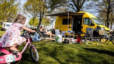 Een foto van een vrouw die in een campingstoel bij een geel kampeerbusje zit te lezen, op de voorgrond een klein meisje met een roze fiets