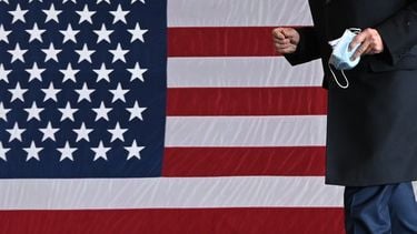 Een foto van de Amerikaanse vlag, Joe Biden staat ervoor met een mondkapje in zijn hand.