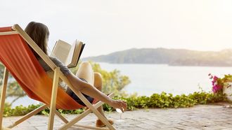Een foto van een vrouw die in een oranje strandstoel een boek leest op een terras aan een meer. Ze heeft een glas wijn in haar hand. Op vakantie.