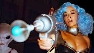 Een foto van Rita Ora met blauw haar een een scifi-wapen, links van haar staat een alien