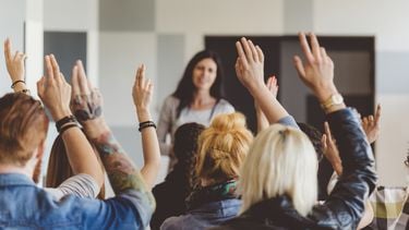 Een foto van een groep zittende mensen die hun hand opsteken, op de achtergrond hun leraar
