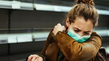 Een foto van een vrouw in een bruin jasje die met mondkapje op in haar ellenboog niest