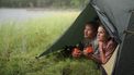 Een foto van een jong stel dat samen uit een tent kijkt op de groene oever van een meer. Ze hebben oranje kampeerbekers vast. Staatsbosbeheer sluit alle wildkampeerterreinen.