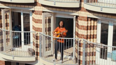 Jeangu Macrooy gitaarspelend op een balkon tijdens Woningsdag.