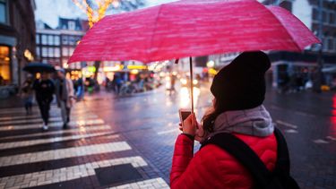 Vrouw met muts op straat onder paraplu met telefoon in de hand