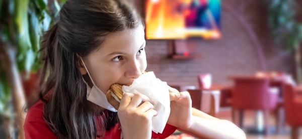 Op deze foto zie je een meisje een tosti eten met haar mondkapje nog om