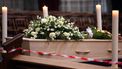 Een foto van een begrafeniskist met bloemen erop, er zit een rood met wit lint voor zodat niemand te dichtbij komt