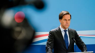 Een foto van een serieus kijkende premier Rutte tegen een blauwe achtergrond, een onscherpe filmcamera links in het beeld.