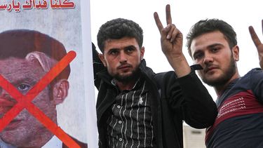 Op deze foto zijn twee mannen te zien met een bord waarop president Macron is afgebeeld met een varkensneus. Er staat een rood kruis over zijn gezicht.
