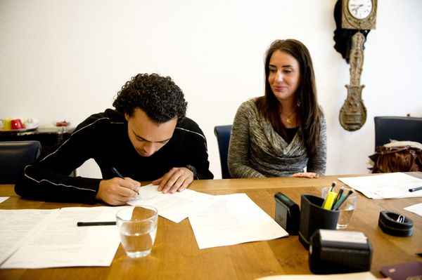 Een foto van een stel aan een tafel, een man zit over papieren gebogen om zijn handtekening te zetten, een vrouw kijkt toe.