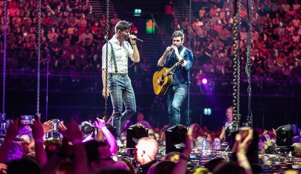 Een foto van Nick en Simon tijdens een optreden in de Ziggo Dome, Nick draagt een blauw shirt en een gitaar, Simon draagt een wit shirt.