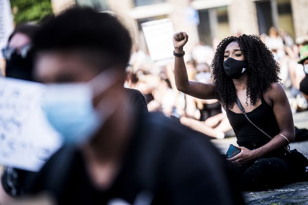 Een foto van een jonge vrouw met mondkapje zittend op de grond, op de voorgrond onscherp een andere demonstrant met mondkapje