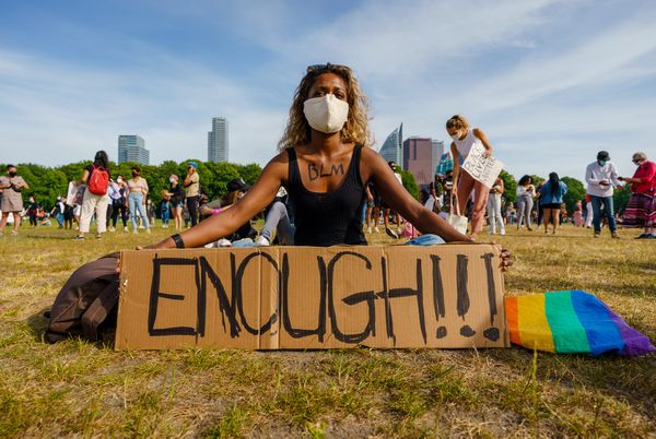 Een foto van een jonge vrouw met mondkapje op het malieveld in den haag, ze zit met een bord waarop 'enough' staat. Op de achtergrond meer demonstranten en de stad Den Haag.