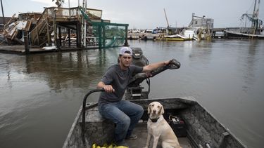 Op deze foto is een man te zien op een bootje met een hond. Ze varen langs verwoeste huizen.